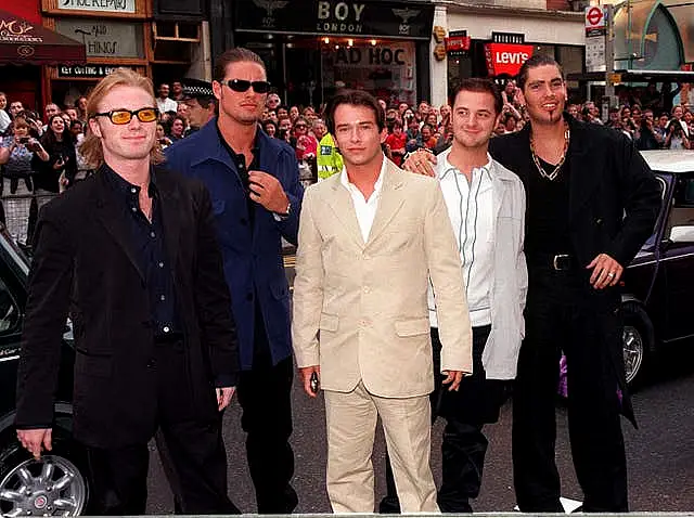 Boyzone line up in a street with cars and fans behind them