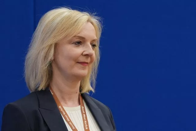 Former UK prime minister Liz Truss wearing a black jacket, a white top, standing in front of a blue background