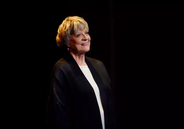 Dame Maggie Smith smiling, wearing a black jacket with a black background