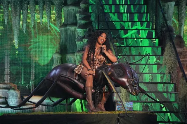 SZA sits astride a large model insect on stage with a green background