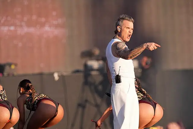 Robbie Williams points out to the crowd while female dancers behind him stick out their bottoms