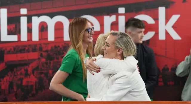 Two women in white hug in front of a red sign that says 'Limerick'. A red-haired lady in a green shirt walks past in the background