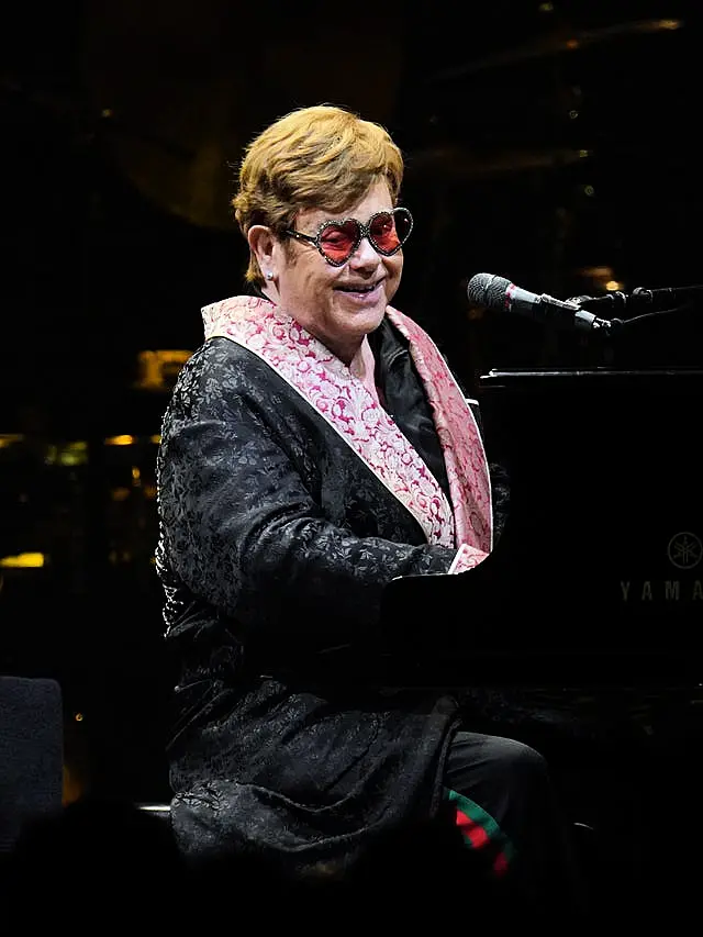 Sir Elton John smiling in heart-shaped sunglasses while playing the piano