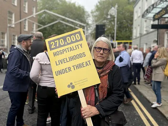 A woman with blonde hair and glasses holds a yellow placard that reads '270,000 hospitality livelihoods under threat'