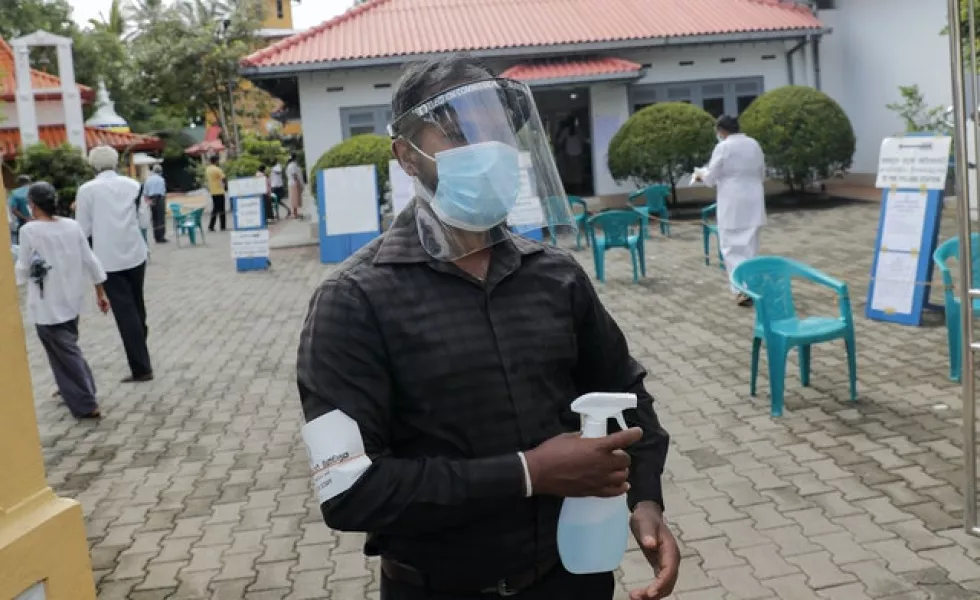 A Sri Lankan polling officer wearing mask and face shield stands holding a sanitiser sprayer (Eranga Jayawardena/AP)