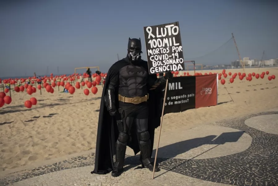 A man dressed in a Batman costume holds a sign with a message that reads in Portuguese ‘Mourning 100,000 deaths of Covid-19, Bolsonaro genocide’ (Silvia Izquierdo/AP)
