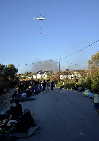 Migrants gather outside the camp as a helicopter tackles the fire (Panagiotis Balaskas/AP)