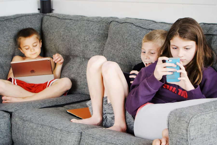 Three children sit on a couch with phone and tablets.