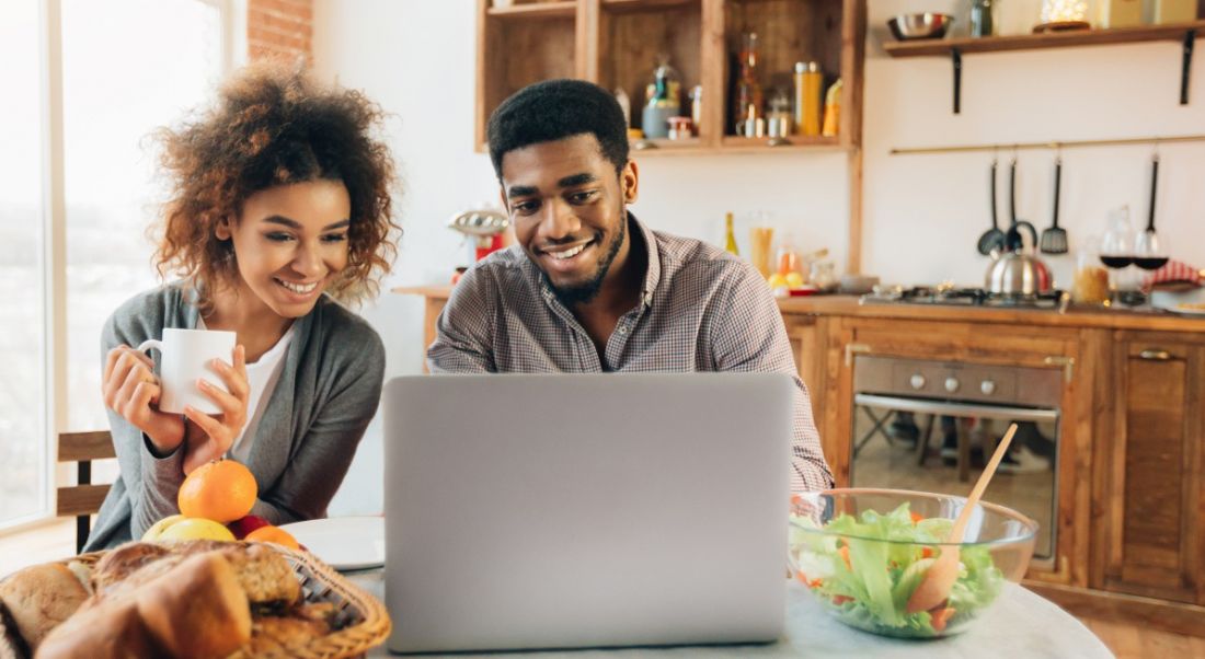 People sitting in oak wood kitchen crowded around silver laptop looking at the screen and smiling.