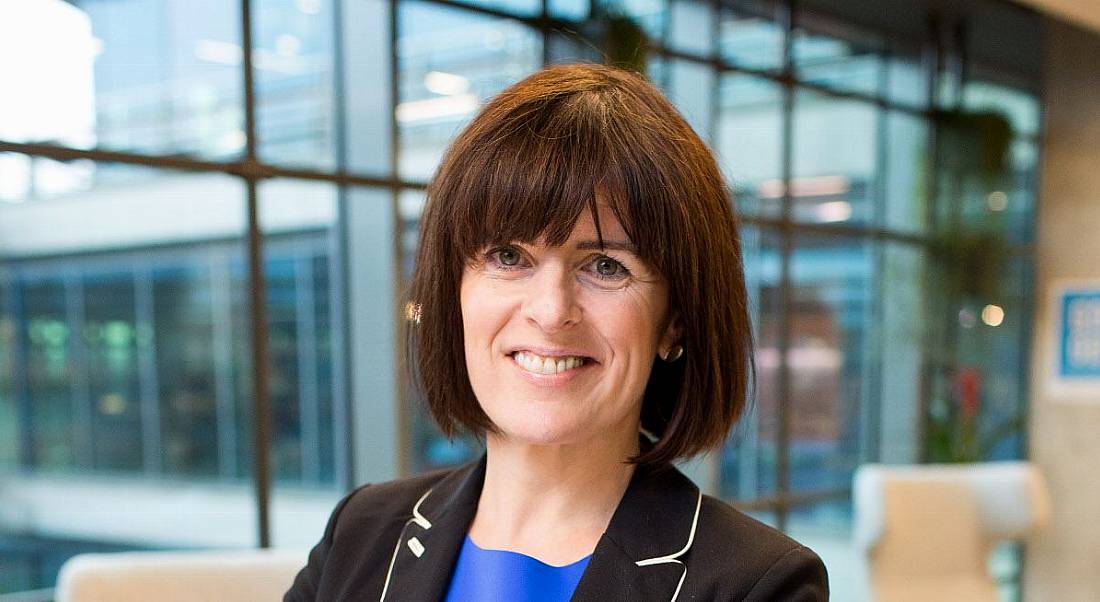 Adrienne Gormley, a woman with short brown hair wearing a black blazer, smiles at the camera. There are large office windows behind her.