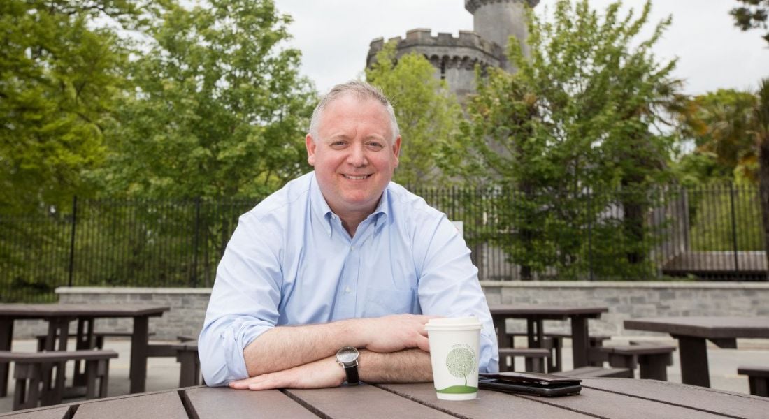 Niall O'Leary sits outside having a coffee at a wooden table, a castle tower visible in the background