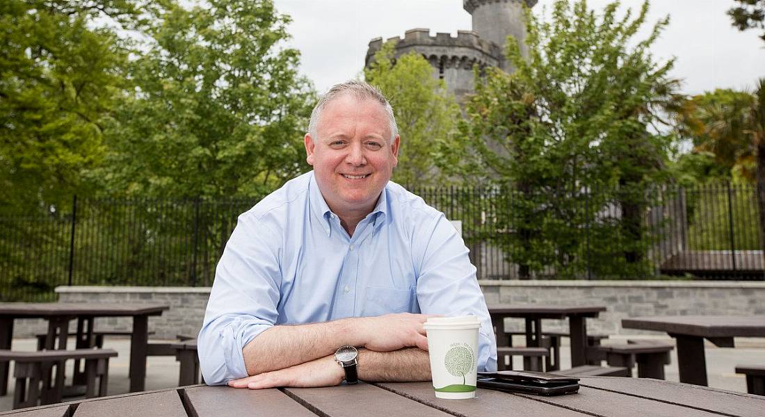 Niall O’Leary pictured having a coffee outside at a wooden table, a castle tower visible among the trees in the background