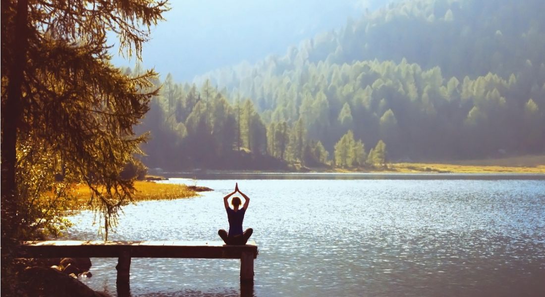 Wellbeing concept depicted by a woman sitting on a jetty in a yoga position facing a breath-taking lake and forest.