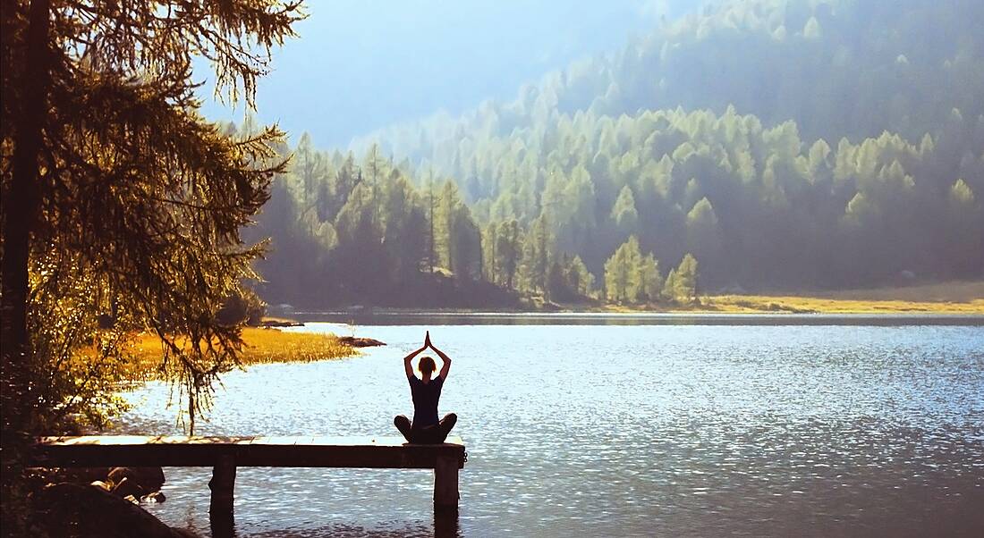 Wellbeing concept depicted by a woman sitting on a jetty in a yoga position facing a breath-taking lake and forest.