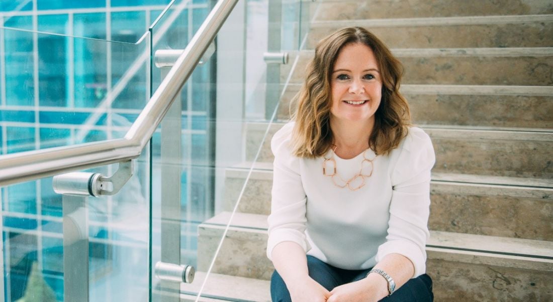 Dr Alice Kirby smiles while sitting on a staircase next to a window looking out at a glass-fronted building