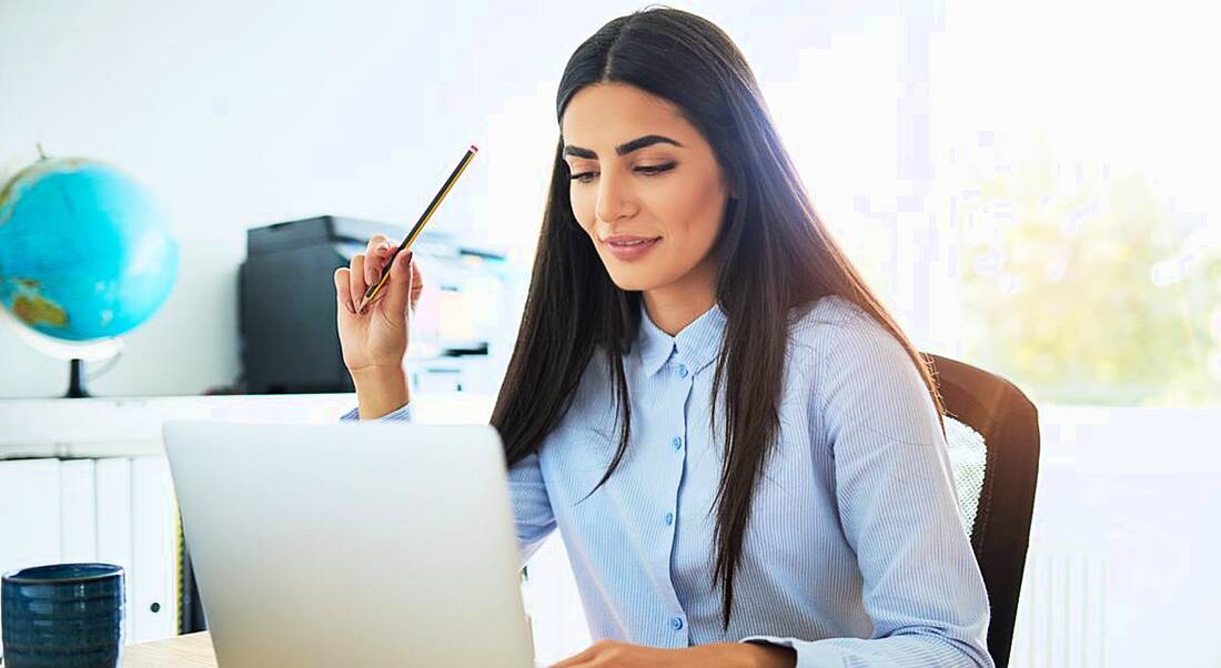 Young woman returning to work, sitting at a desk in front of a laptop holding a pencil.