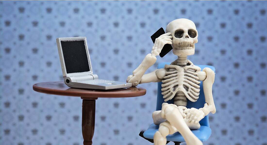 Toy skeleton sitting at a desk with a laptop, talking on a smartphone to depict overworking.