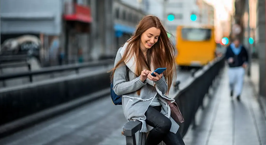 Woman sitting on a rail in a city, smiling and texting her friends to stay in touch.