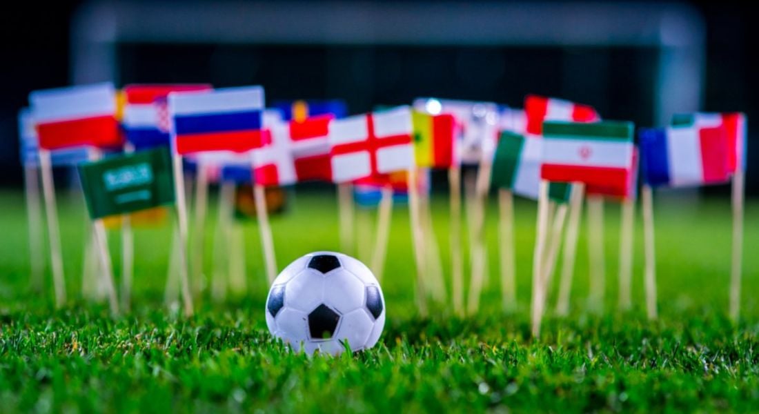 A soccer ball sitting on the grass in front of various miniature flags depicting the World Cup