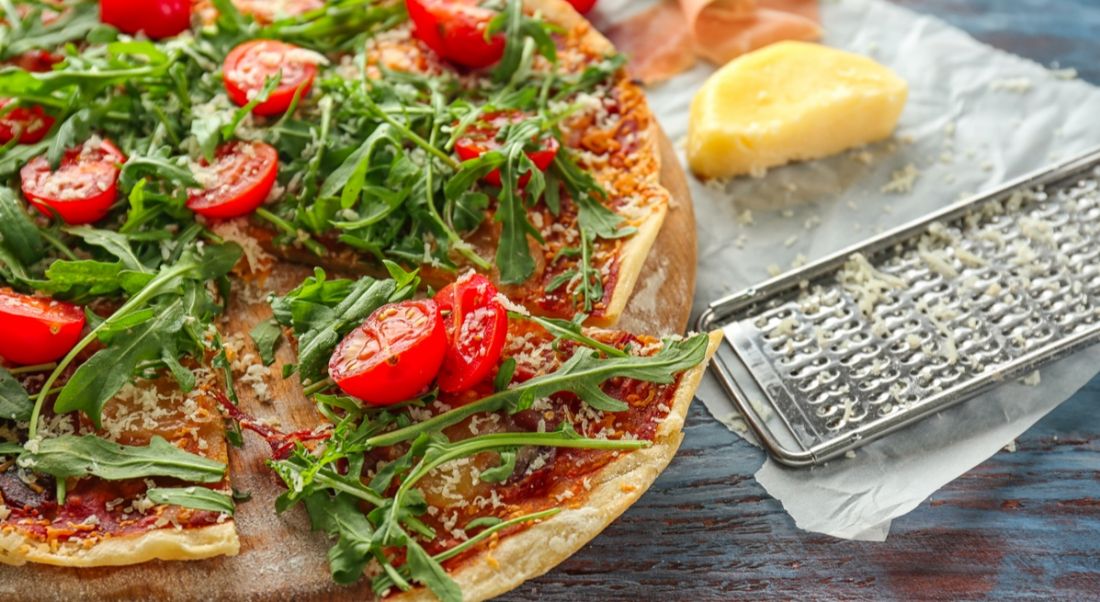 Pizza covered with tomasto slices, rocket and serrano ham on top of wooden board. Slice pizza company