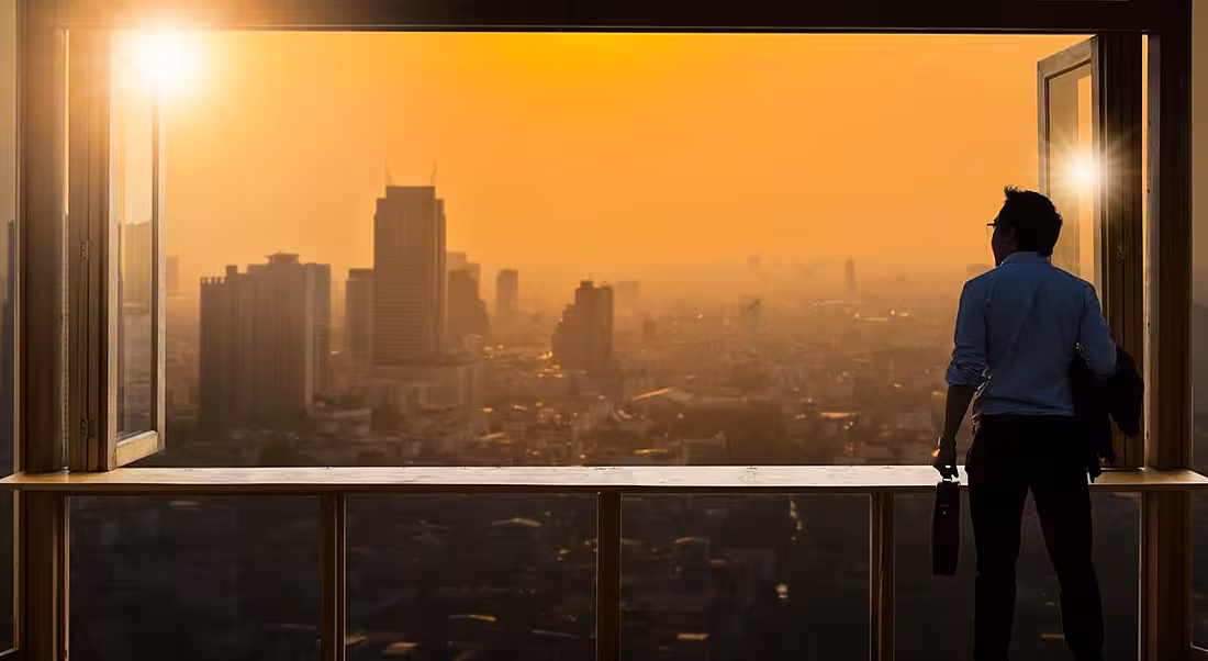Young business man looking out window at sunrise in metropolitan area. Looking ahead at trends.