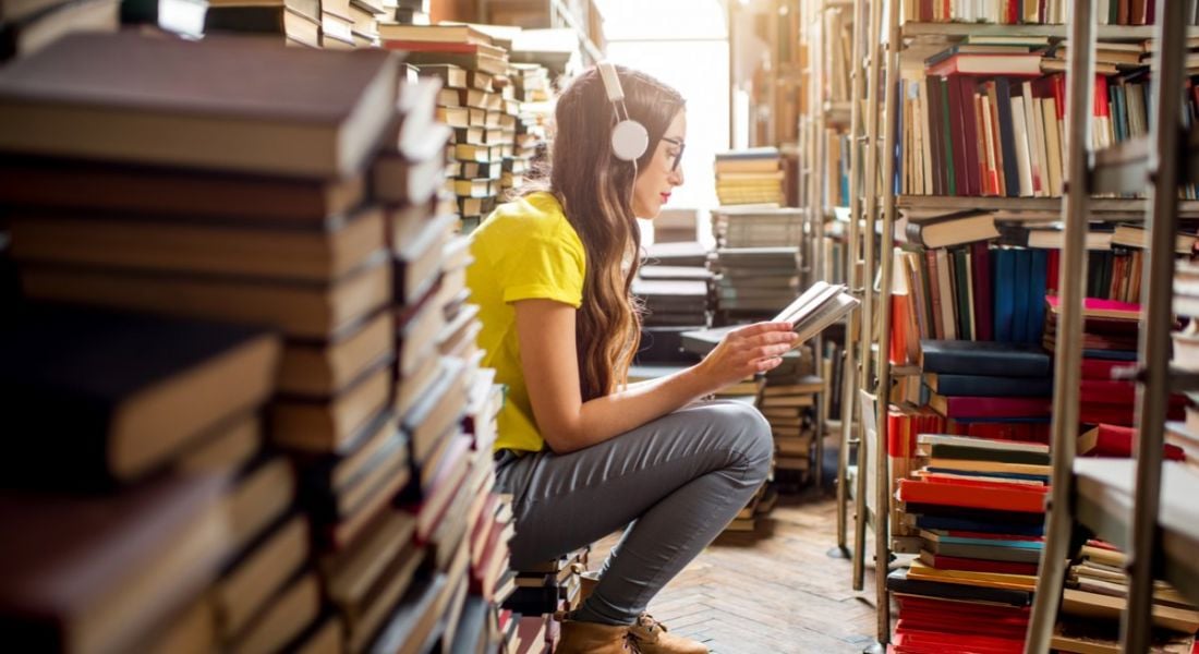 Young woman sitting in a library reading with headphones on surrounded by books trying to reskill for her career.