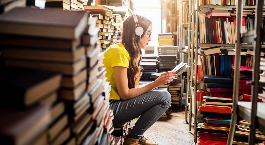 Young woman sitting in a library reading with headphones on surrounded by books trying to reskill for her career.