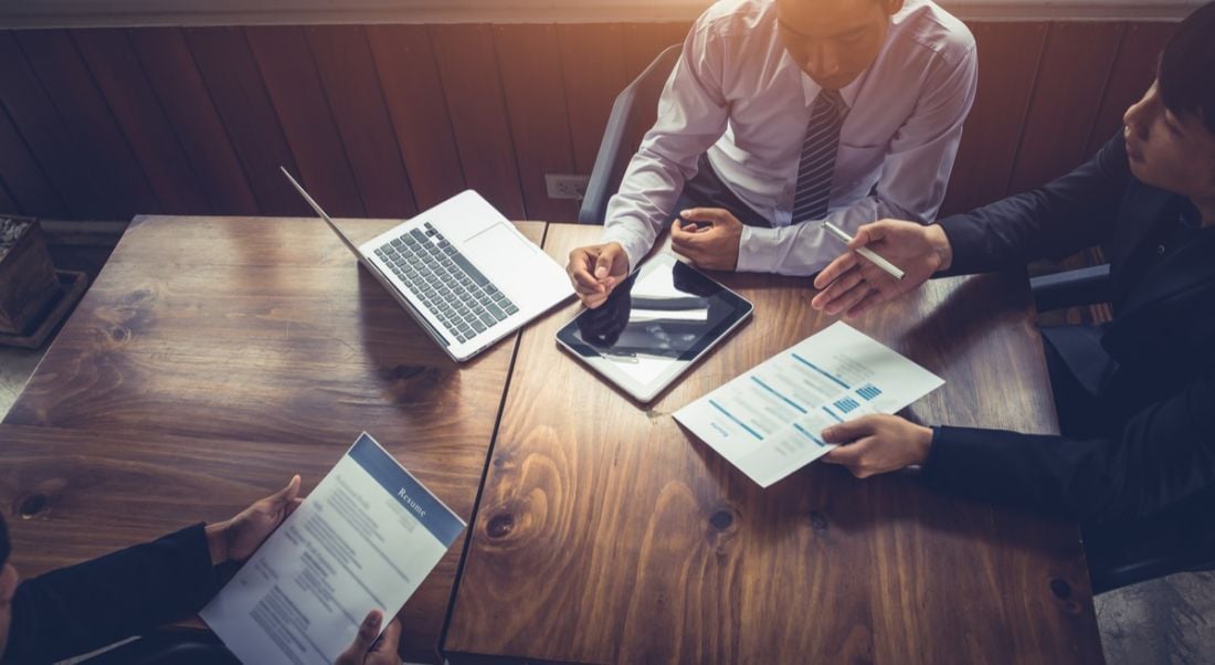 Businessmen at a mahogany table holding a job interview using laptop, iPad, and papers. Jobseekers need to be aware of these emerging industry trends