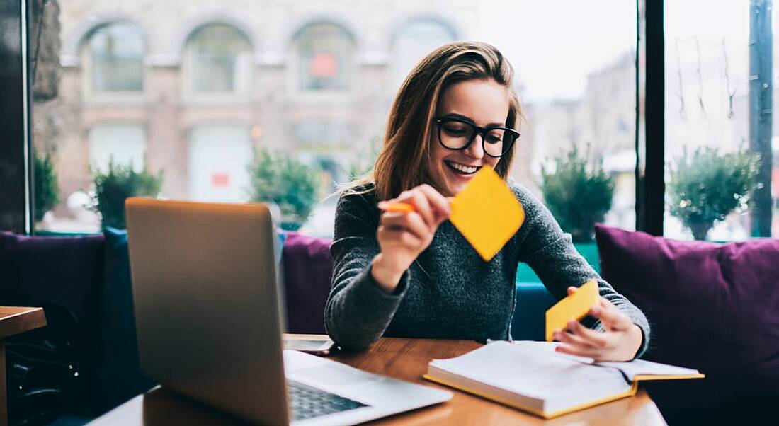 Young woman sitting at a laptop and smiling while using sticky notes, using good habits to achieve career success