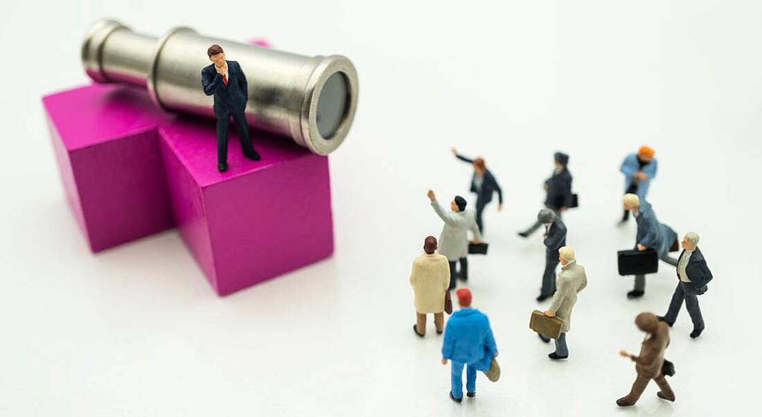 A miniature figure of a man in a suit stands on a pink block beside a telescope observing a small gathering of miniature figures below