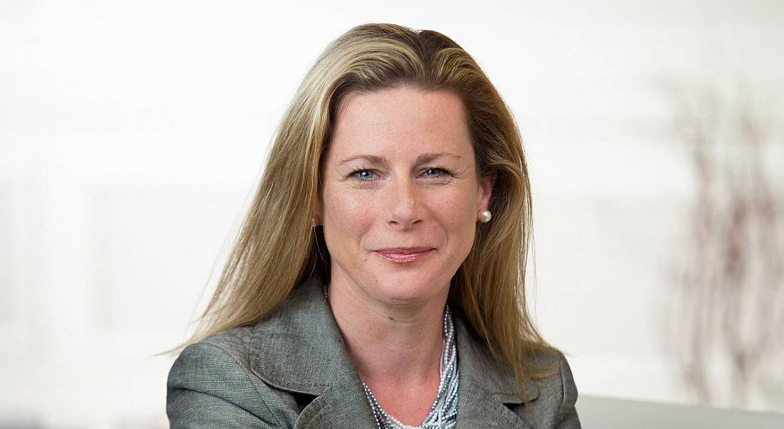 A blonde woman wearing grey jacket and pearls, smiling against white background.