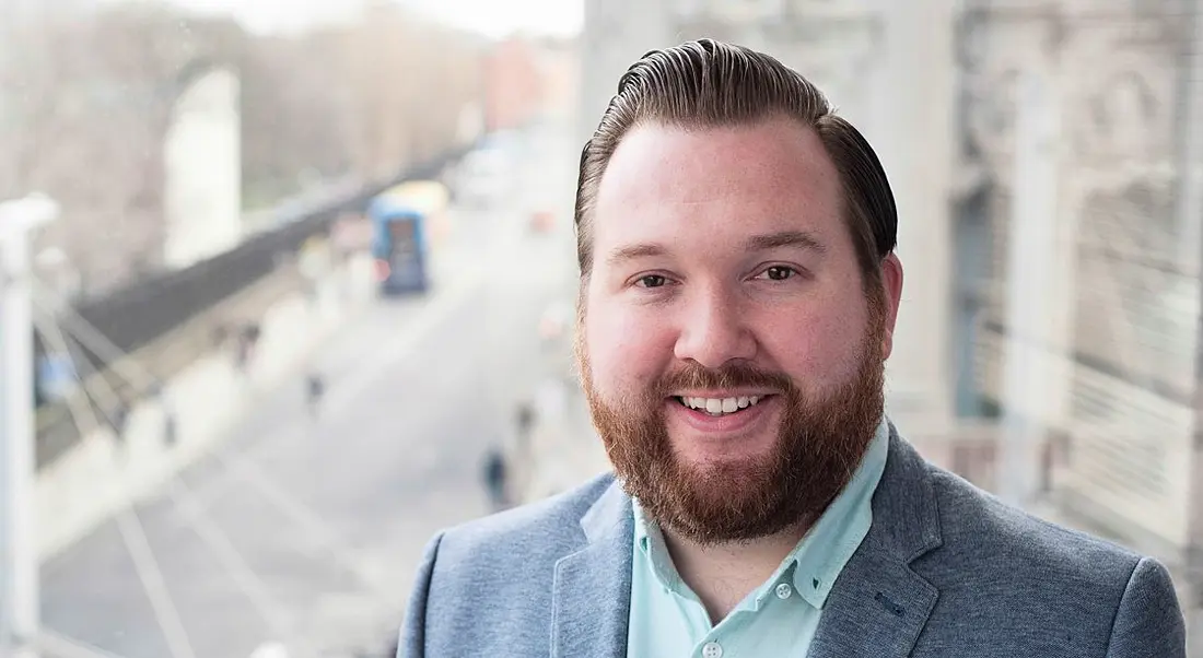 Bearded, red-haired young man wearing a grey suit jacket and blue shirt smiling against an out-of-focus Dublin city backdrop.