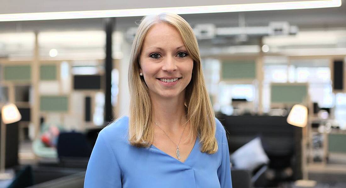 Blonde smiling woman in a light blue shirt smiling with the New Relic open plan office behind her.