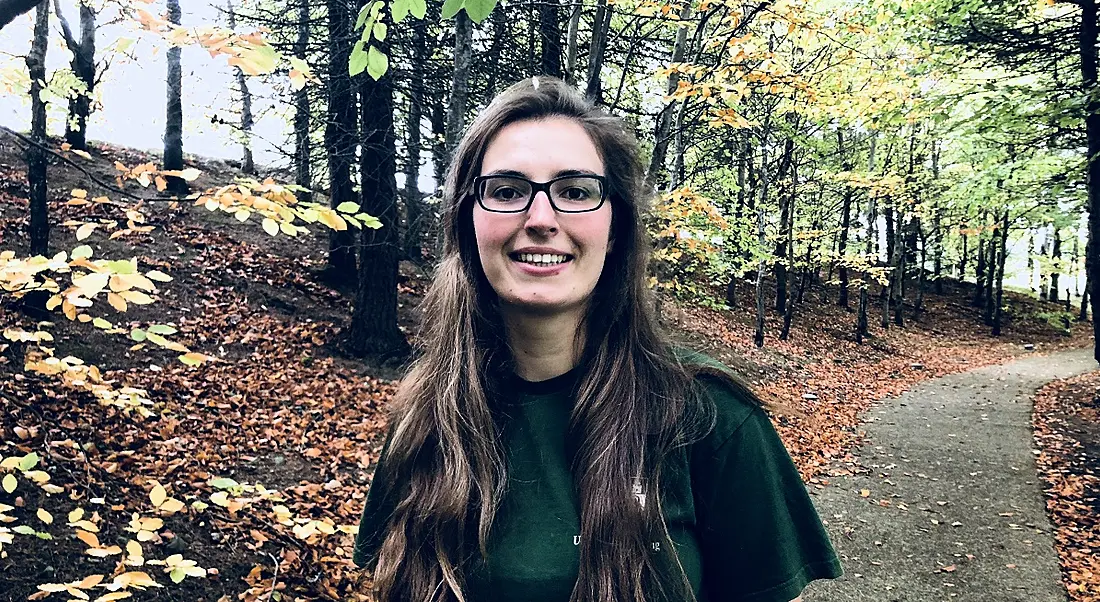 A young woman with long brown hair and glasses standing against the backdrop of a forest during autumn.