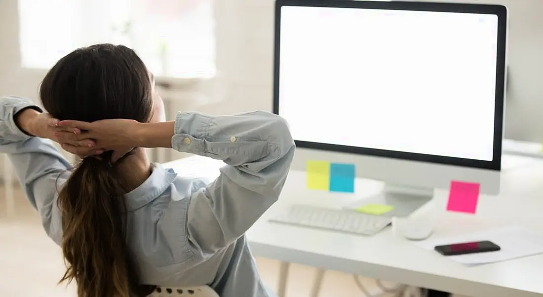 A woman leaning back in her chair in front of her screen with Post-Its on it. She’s taking a break after feeling overworked.