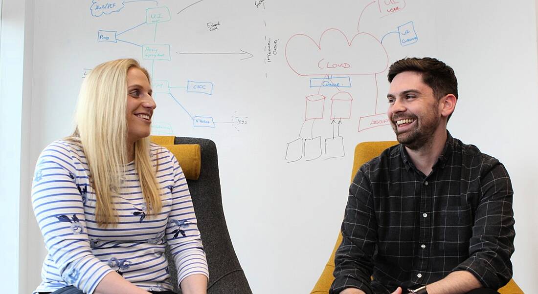 A young blonde woman and a young dark-haired man sitting in front of a whiteboard, laughing and discussing whiteboarding.