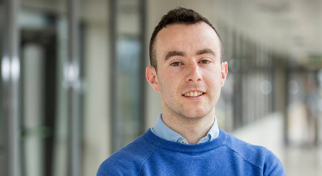 Headshot of a young, dark-haired male graduate who works at Amgen in a blue jumper against a blurred-out building.