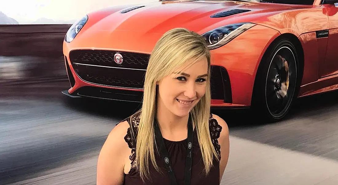 A young blonde woman smiling at the camera against wallpaper depicting a speeding sports car.