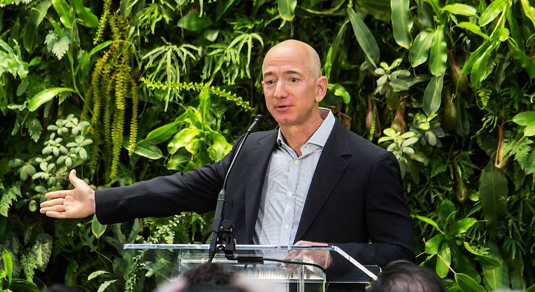 Slim, bald man in his 50s standing at a glass podium in front of a leafy backdrop, gesturing to the side with his right hand.