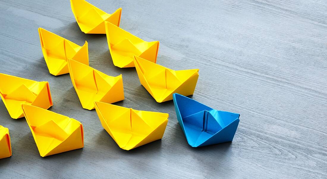 A blue paper sailboat leading a bunch of yellow paper sailboats on a wooden surface to represent leadership skills.