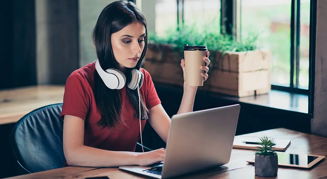 Young woman holding a coffee with headphones around her neck working on a laptop during the quiet season at work.