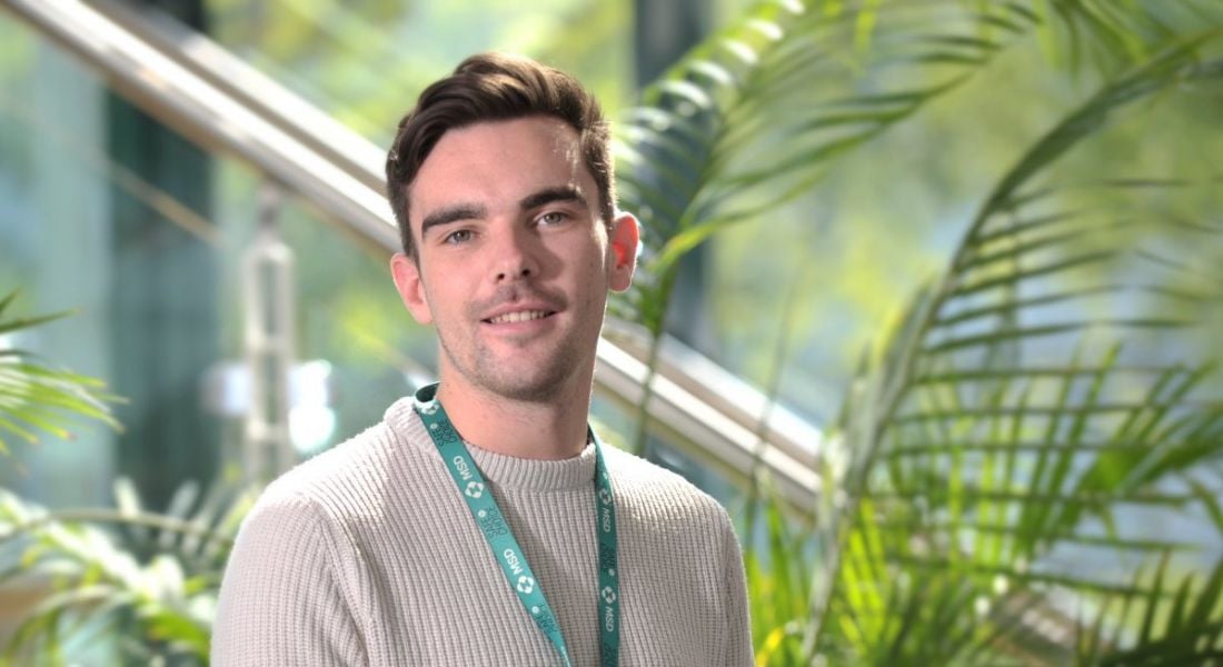 Young male graduate in a beige jumper wearing an MSD lanyard against a backdrop of greenery.