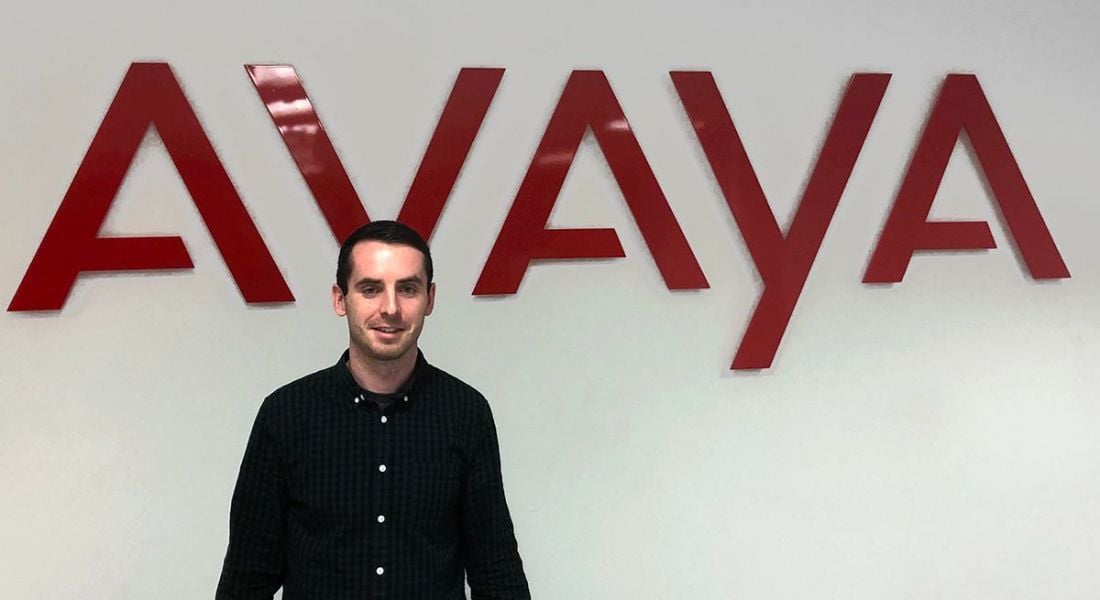 A young, dark-haired man in a black, casual shirt standing in front of a wall that says Avaya in red letters.