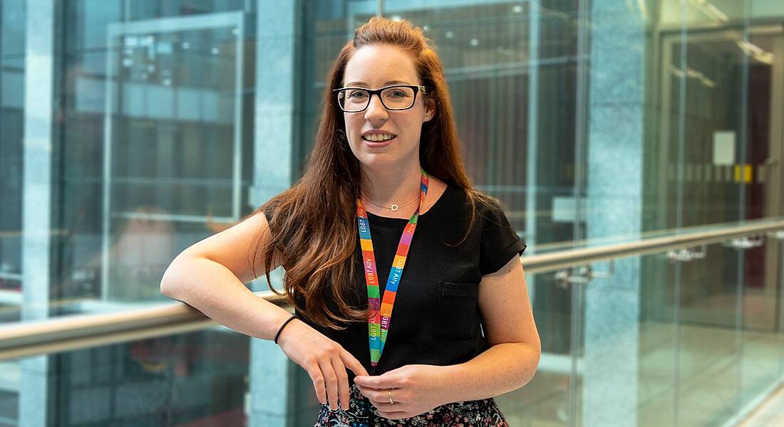 A young woman with cascading auburn hair, blue eyes, thick-framed glasses and a toothy smile leaning against a railing.