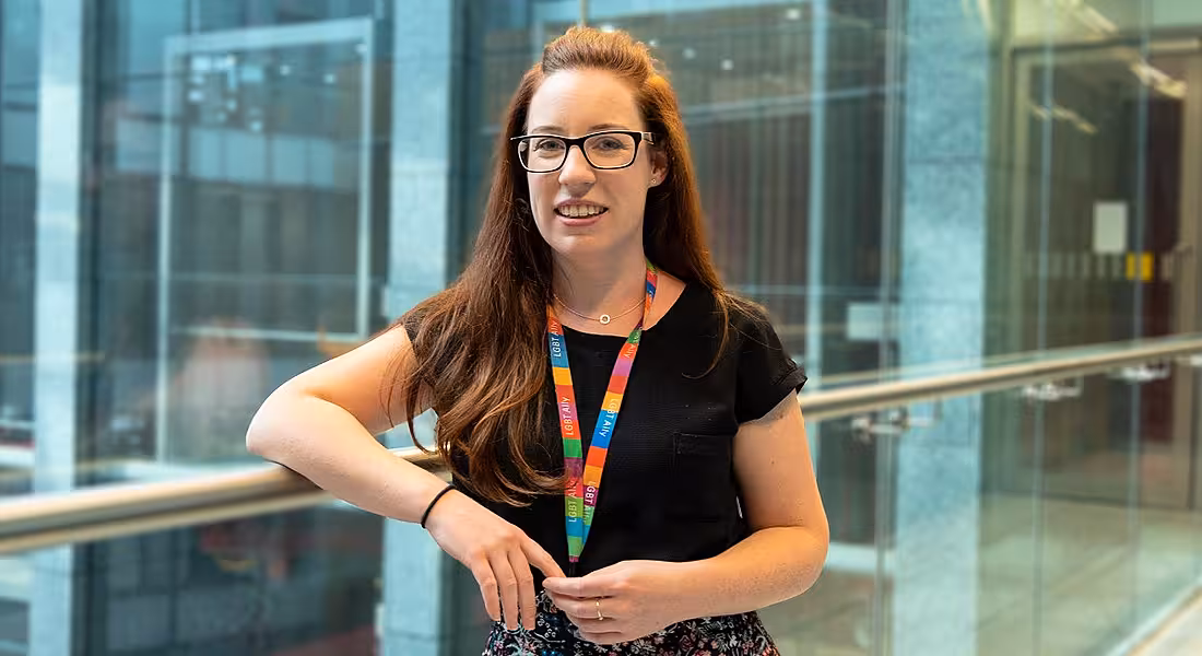A young woman with cascading auburn hair, blue eyes, thick-framed glasses and a toothy smile leaning against a railing.