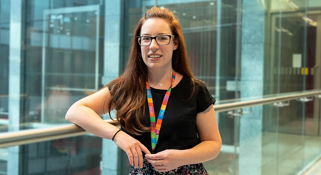 A young woman with cascading auburn hair, blue eyes, thick-framed glasses and a toothy smile leaning against a railing.