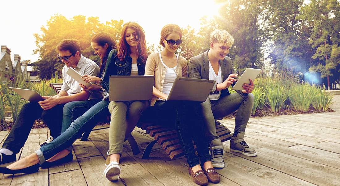 A diverse group of young graduates sitting on outdoor benches looking at their laptops on an overcast summer day.