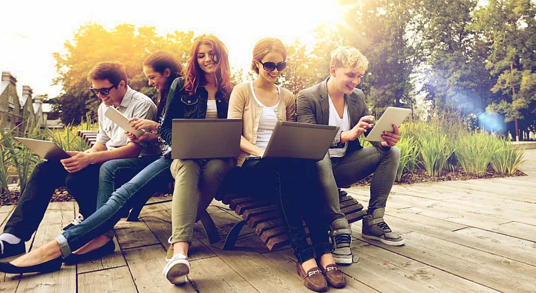 A diverse group of young graduates sitting on outdoor benches looking at their laptops on an overcast summer day.