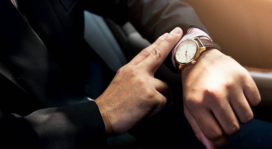 Man in dark suit looking at a watch with a leather strap on his wrist. His face is not in view, only his arm.