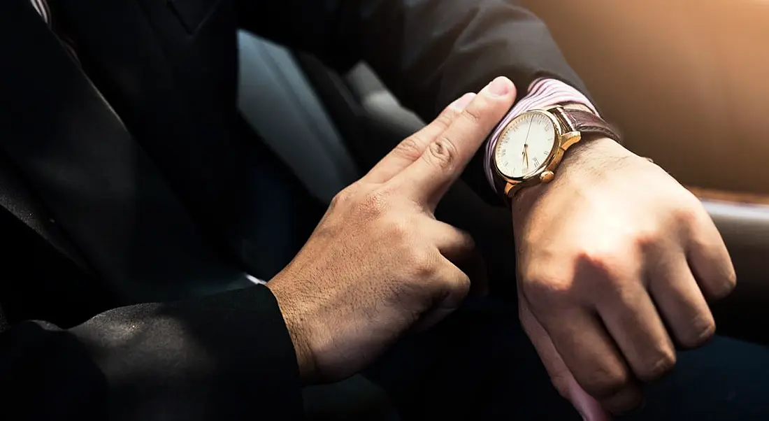 Man in dark suit looking at a watch with a leather strap on his wrist. His face is not in view, only his arm.